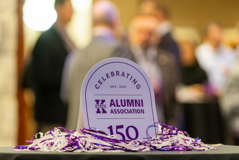 Celebrating 150 years of the K-State Alumni Association lavendar table tent with people gathered in the background